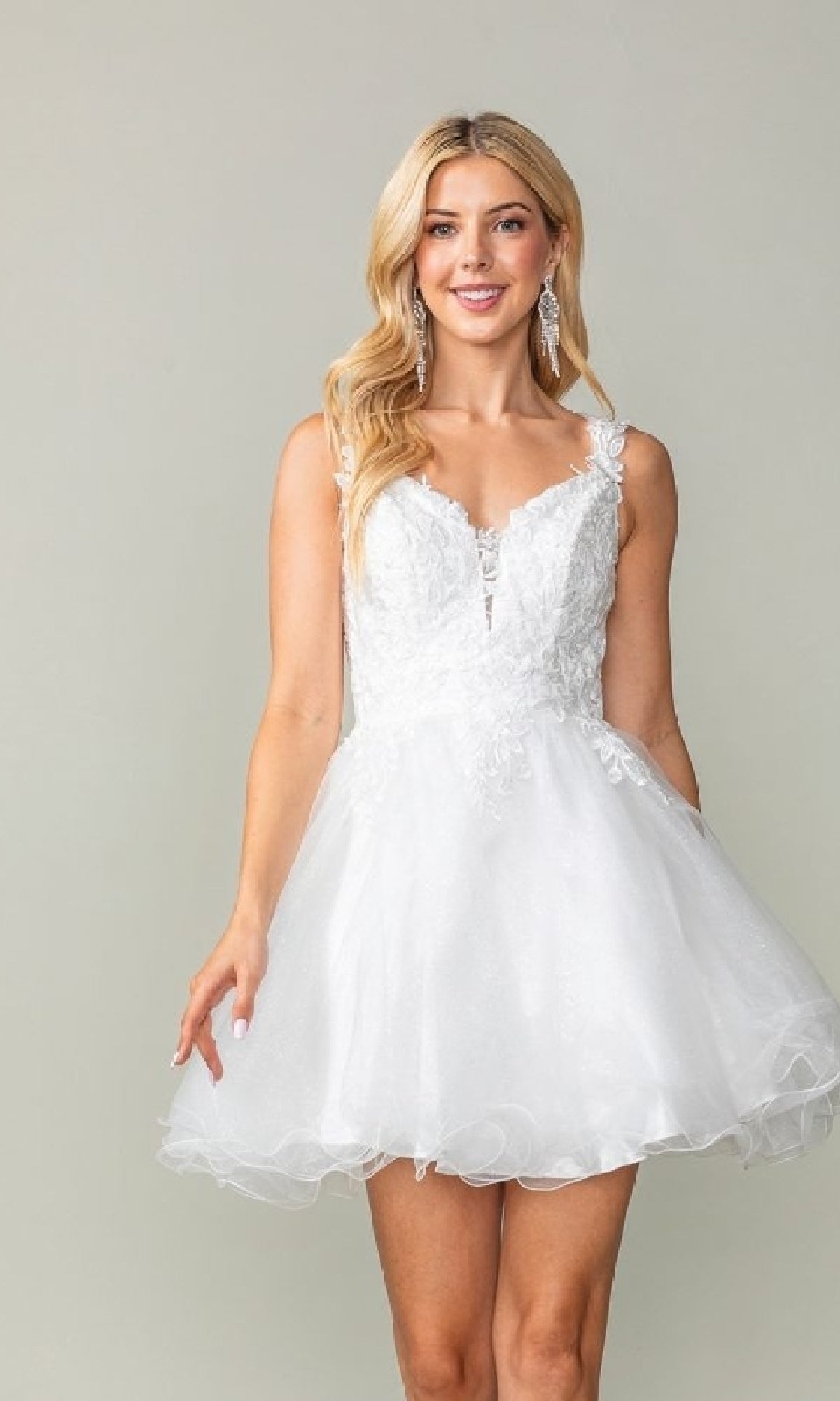 A woman wears the Tulle Babydoll Homecoming Dress: Dancing Queen 3377, a sleeveless, short white lace dress with a flared tulle skirt, standing against a plain background and smiling at the camera.